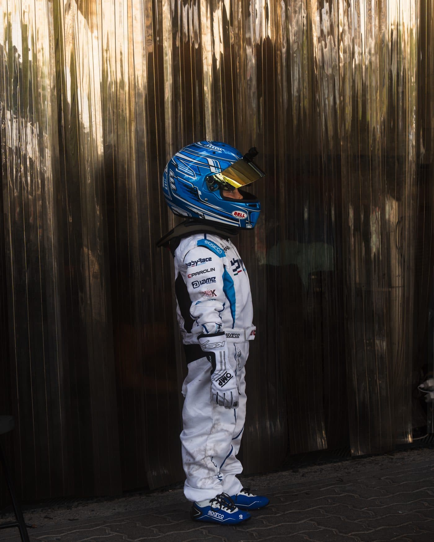 Young driver in full race suit and helmet standing in profile against a chrome curtain
